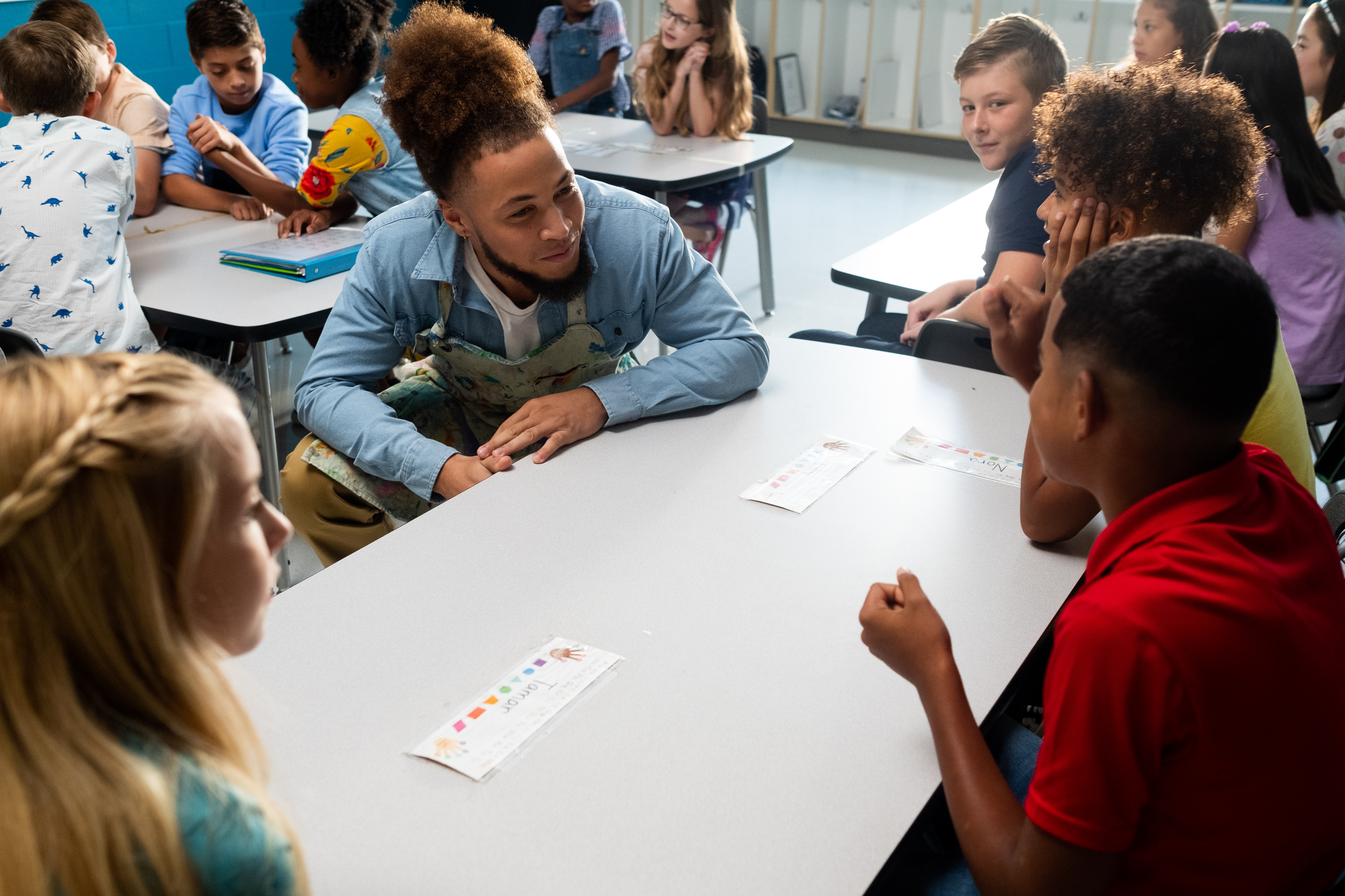 A man in a blue shirt and a group of students sit around a white table talking.