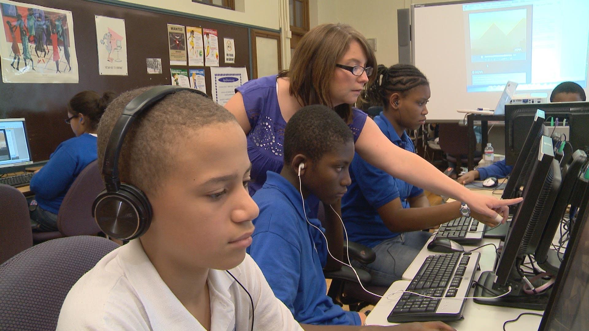 A female teacher points to a male student's computer screen in a computer lab with other students