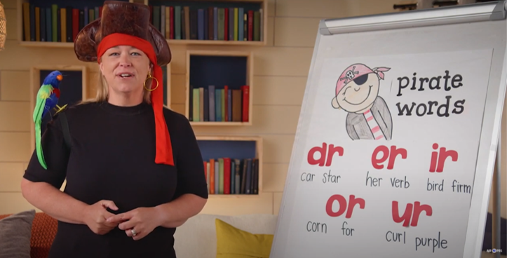 A woman dressed as a pirate next to a white board of vowel sounds.