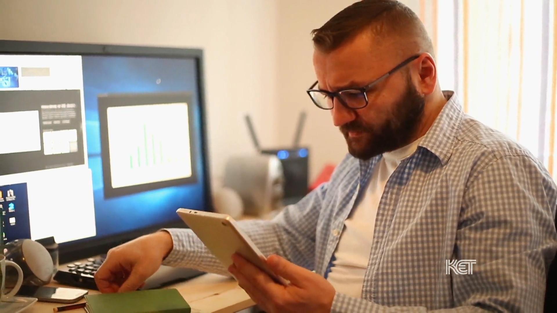 A man holding a tablet and sitting in front of a computer