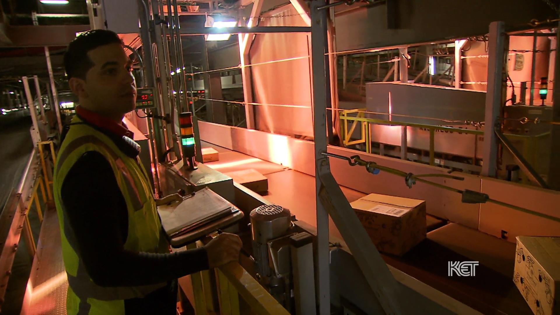 A man in a safety vest with a clipboard monitoring a conveyor belt.