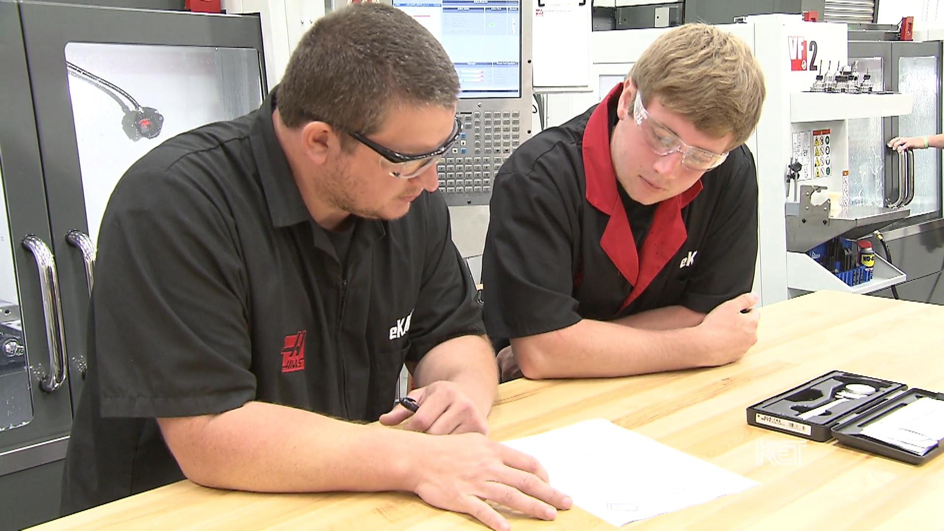 Two men looking at a paper on a work bench