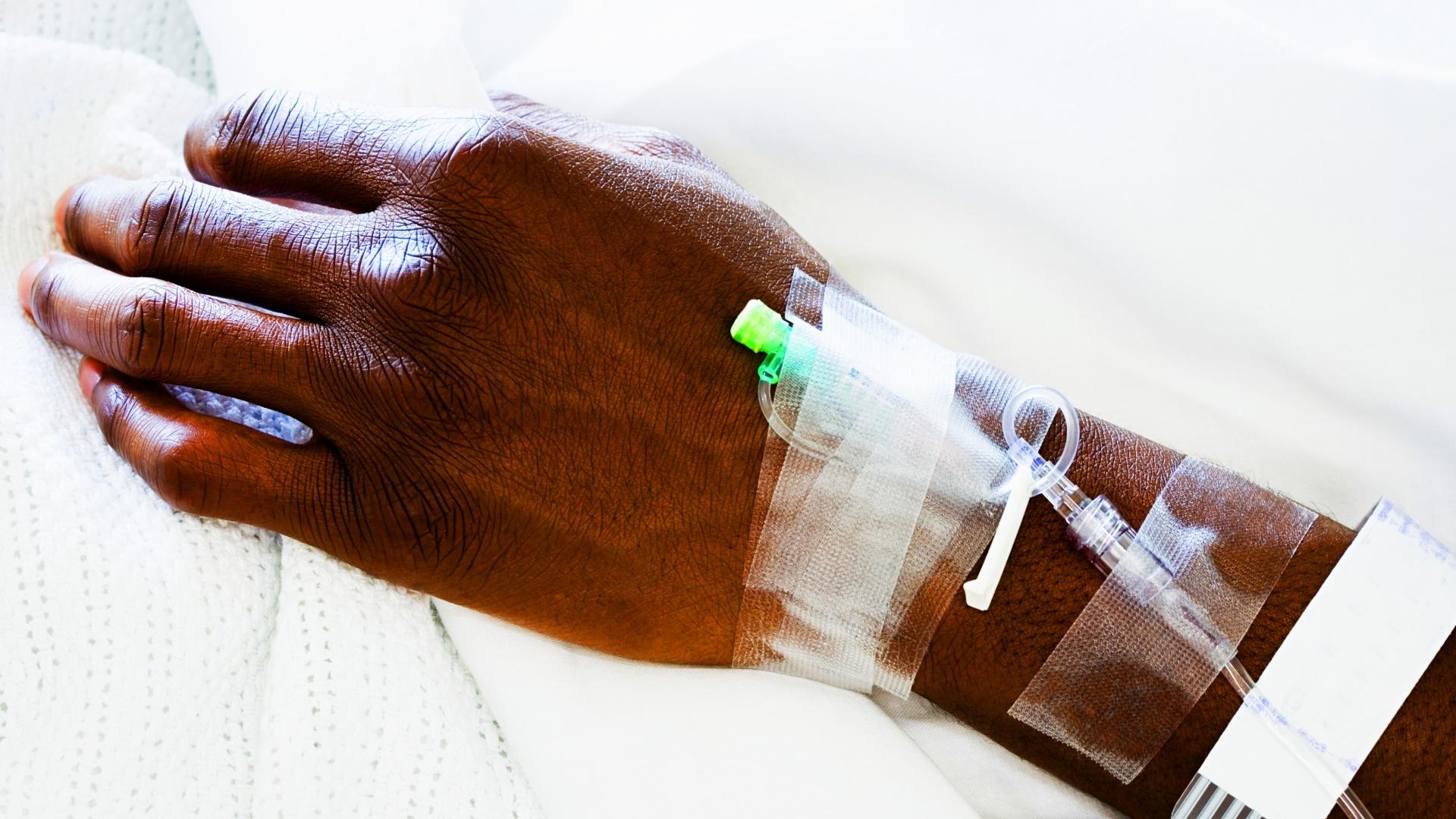 A hand on a hospital bed with an IV taped to it.