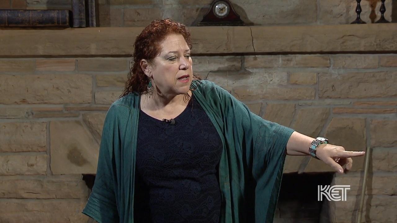 A woman standing in front of a stone fireplace pointing her finger out.