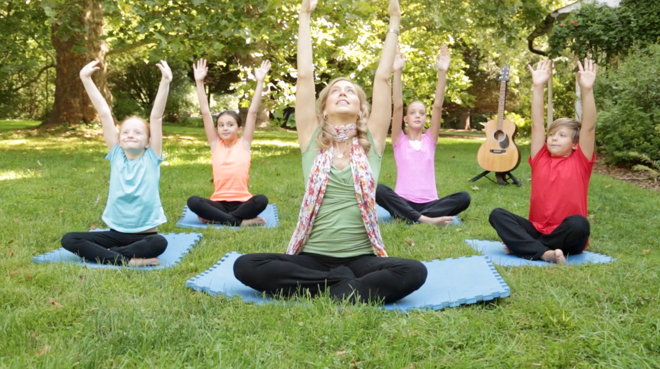 A woman with a yoga class of kids reach their hands up towards the sky.
