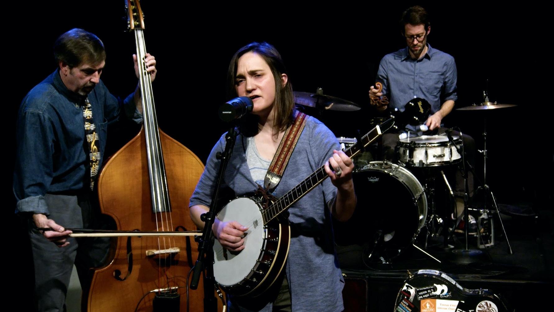 Woman playing banjo and singing into a microphone with man playing an upright bass and drummer in the background.