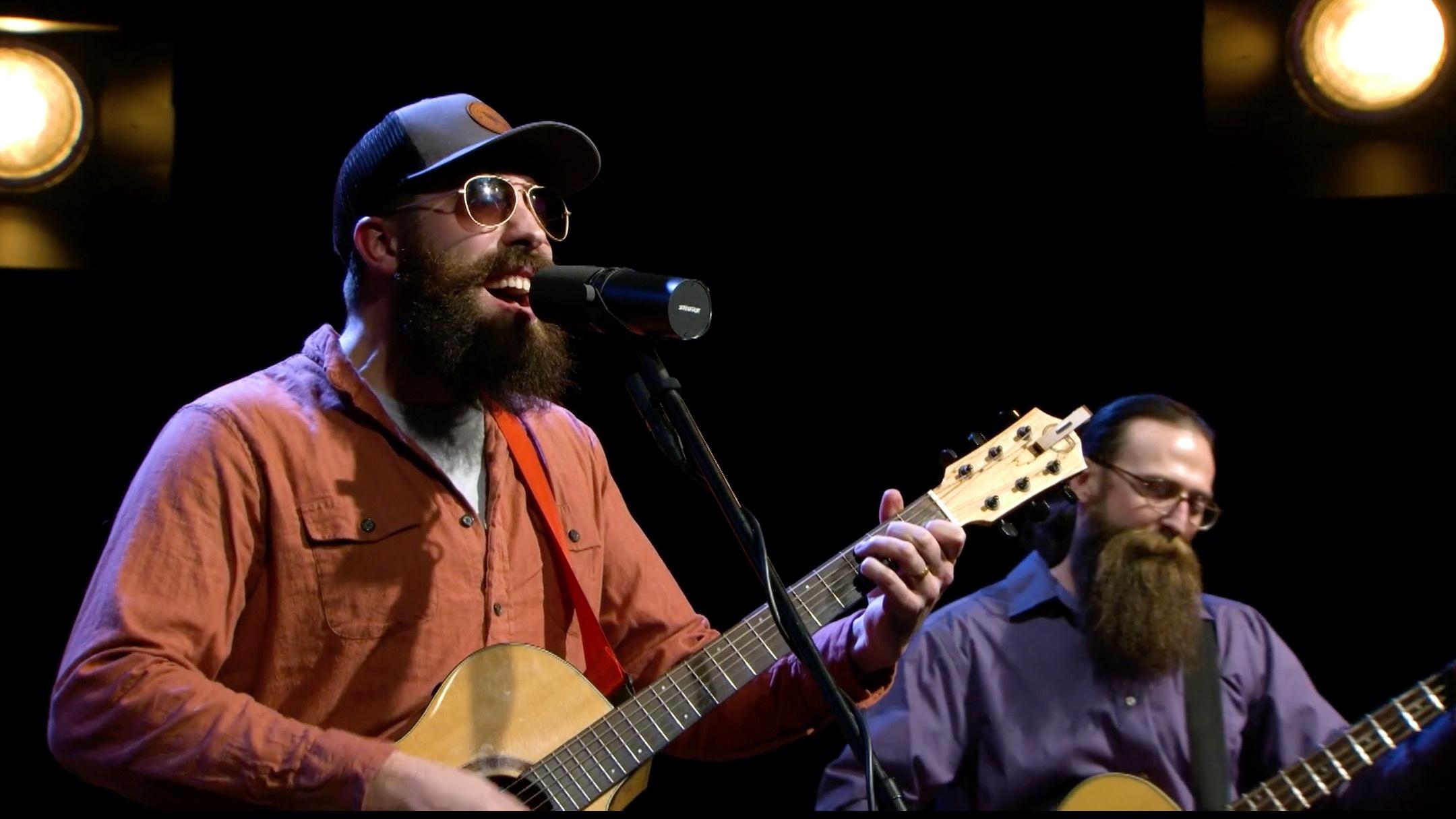 Man with bear, ball cap and sunglasses playing an acoustic guitar while singing into a microphone along with another man with a long beard playing guitar in the background.