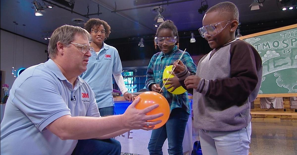 A man holds a balloon while a boy pops it. A woman and girl stand behind them.