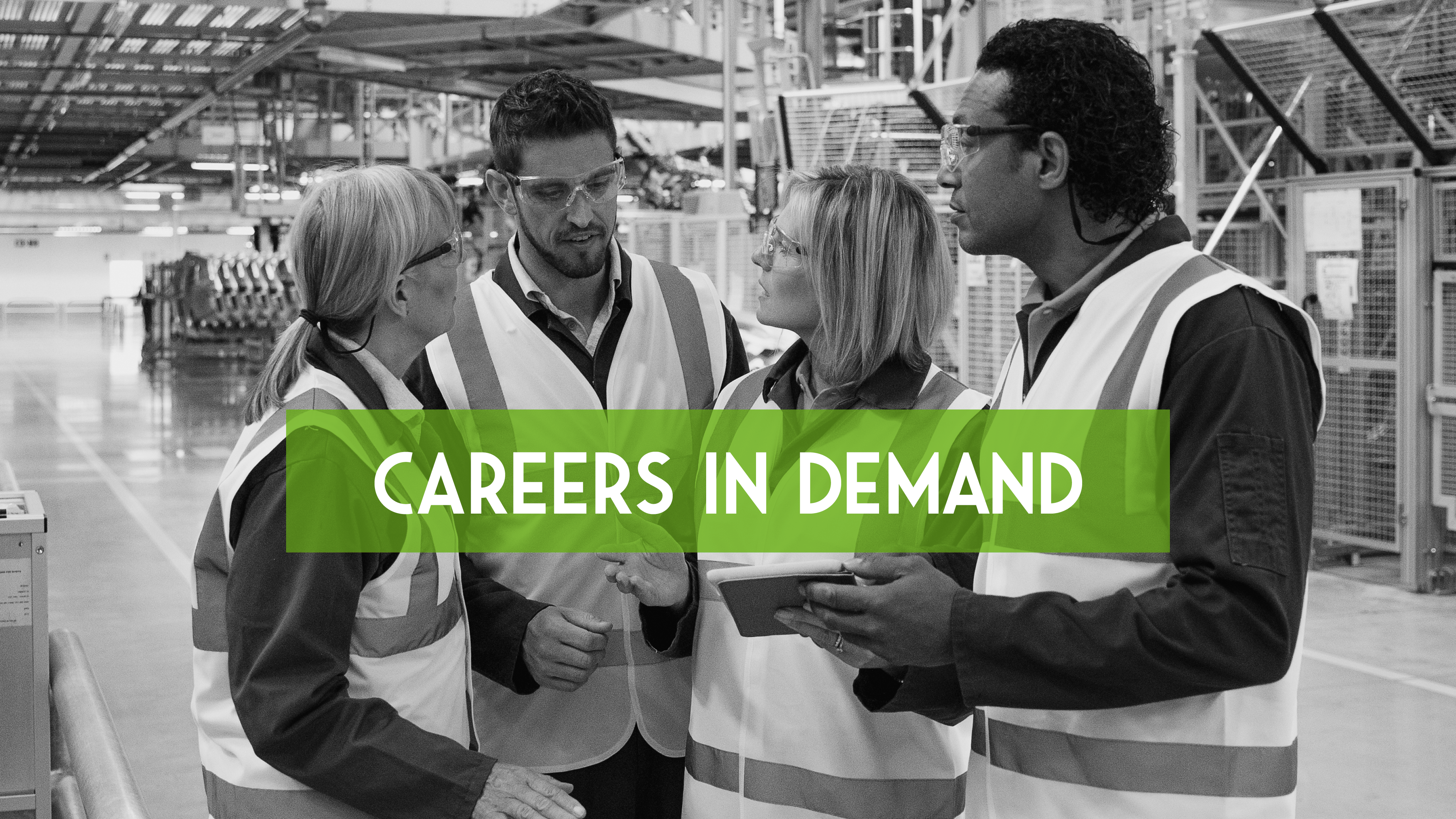 In black and white, two men and two women in safety vests stand together talking in a factory.