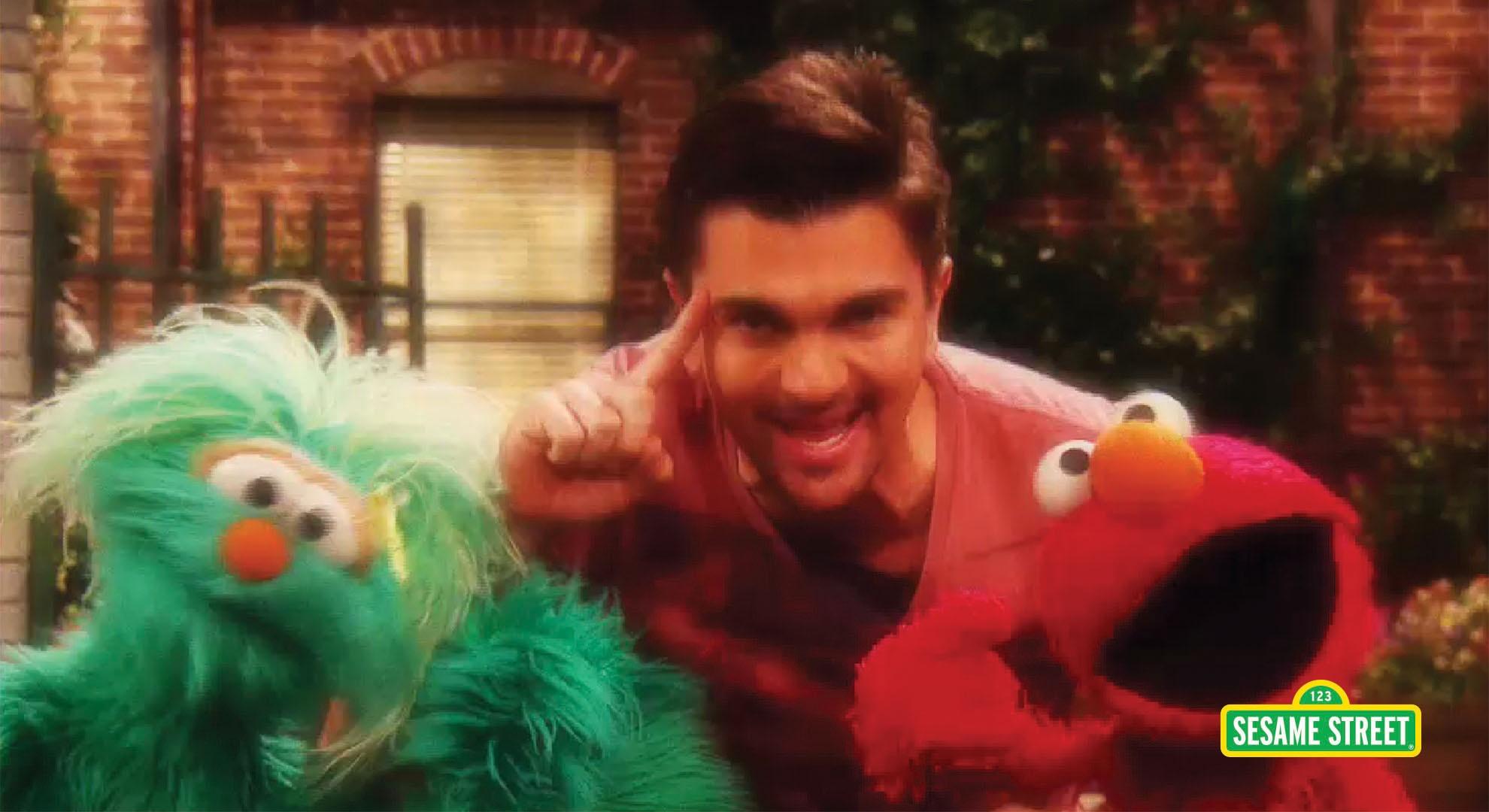 A man puts a finger against his temple and sings with blue and red puppets.