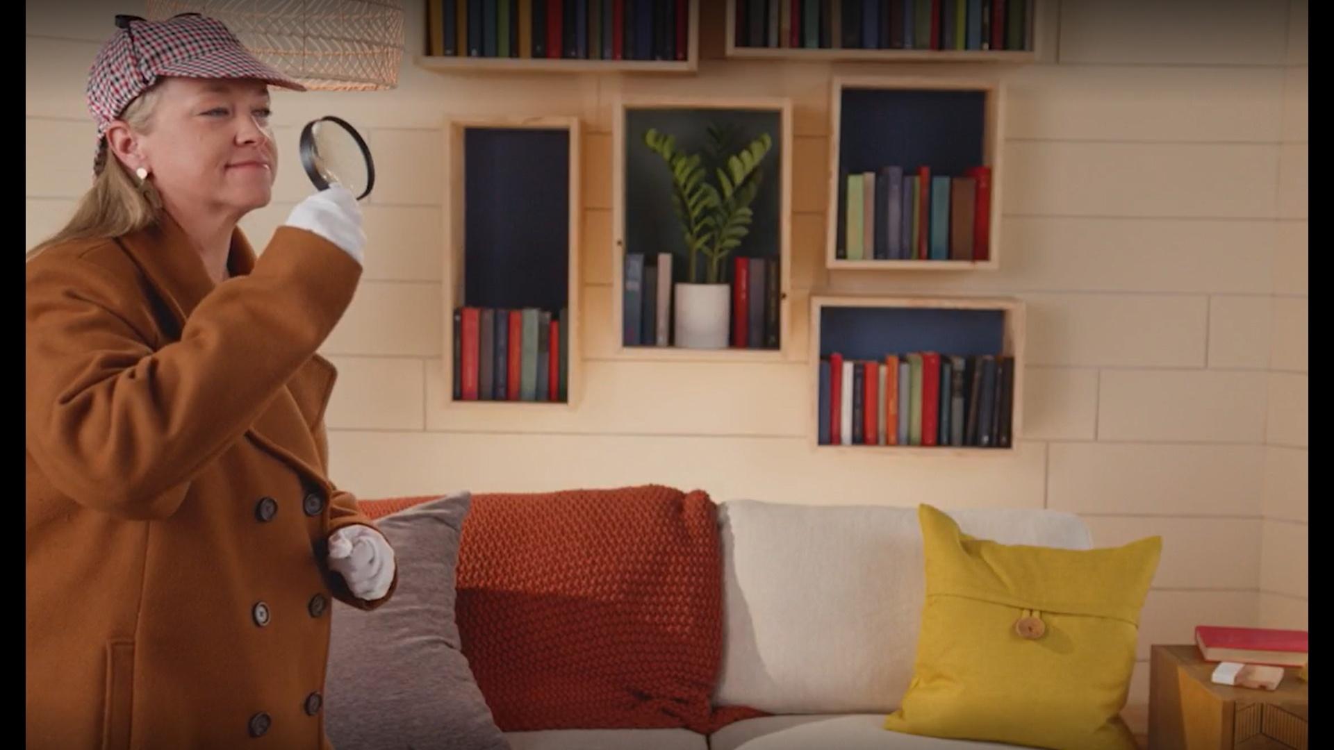 A woman in Sherlock Holmes gear with a magnifying glass stands in front of a sofa.
