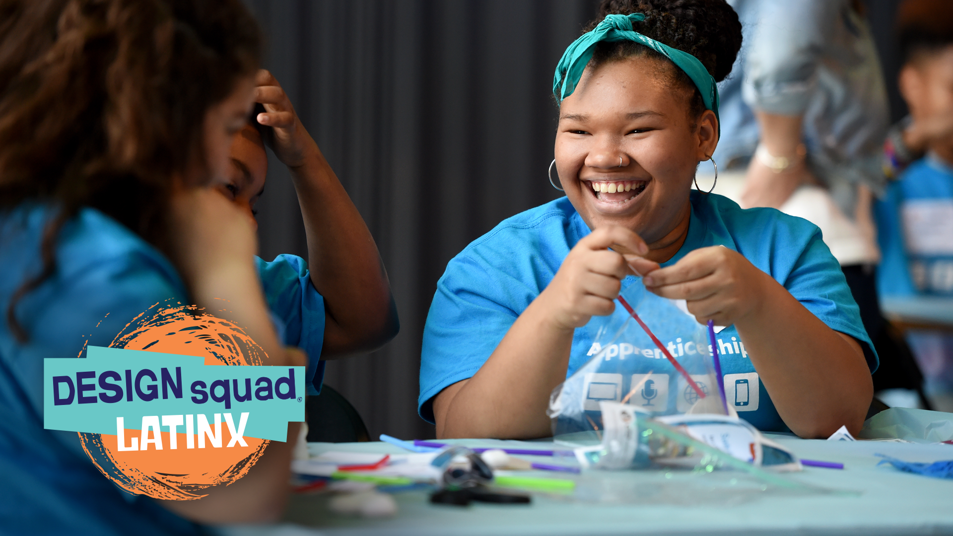 One Black girl smiles at another girl at her table while another child sits between them.
