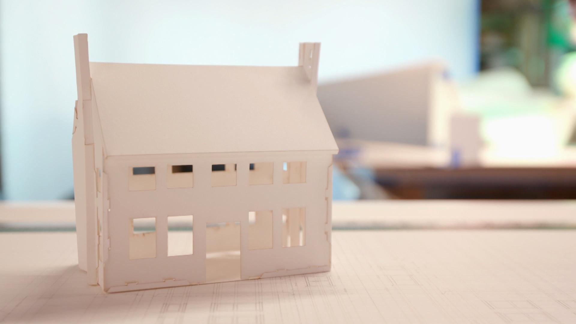 A house made of white paper sits on top of a desk against a blurred background.