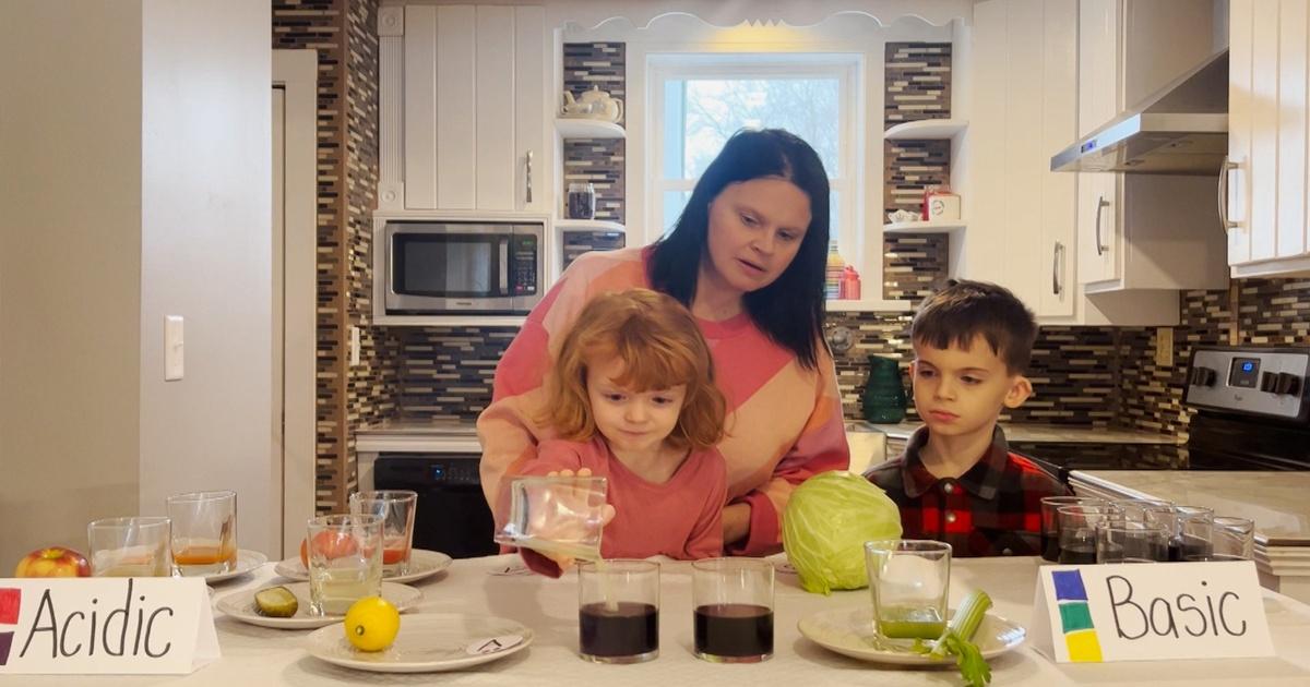 A woman and two children stand behind a counter. The little girl pour a clear liquid into a dark liquid.