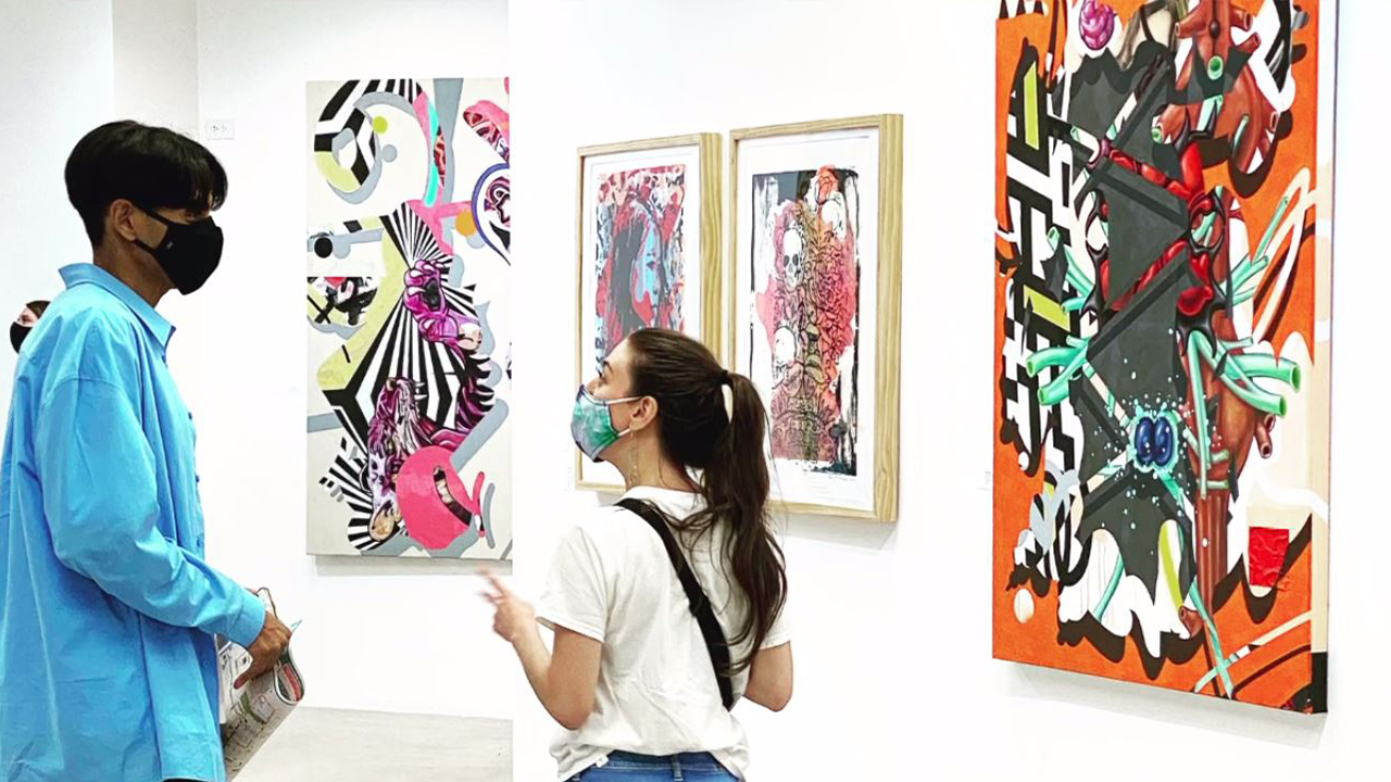 A man in a blue shirt and a woman in a white shirt stand in front of artwork talking.