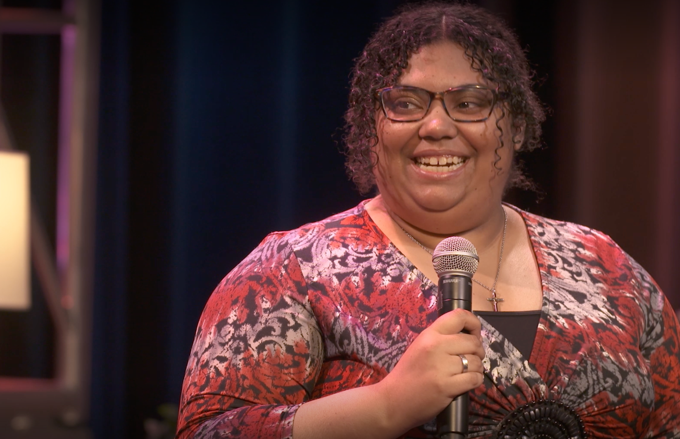 A Black woman with glasses in a printed red top holds a microphone.