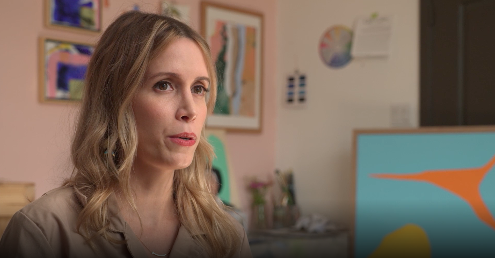A white woman in a khaki top sits in front of paintings and a pink background.