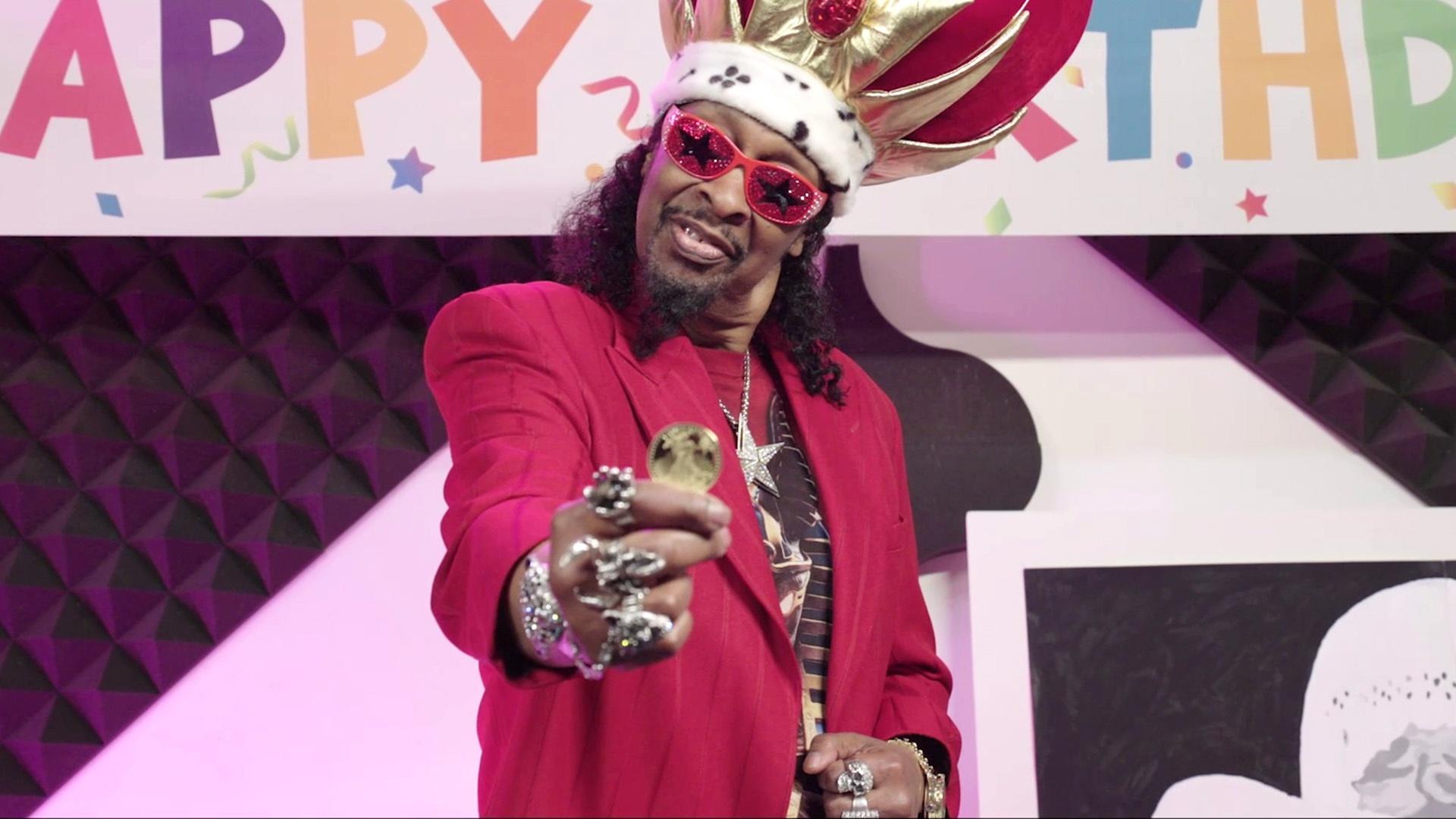 A Black man in a red suit holds a gold coin towards the camera.