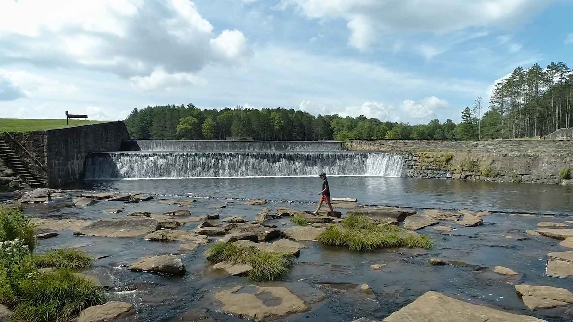 Person walking along rocks across a river in front of a short waterfall.