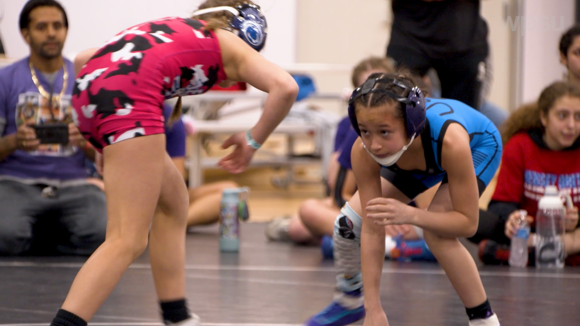 young girl in red singlet with back facing engaging in wrestling posture with another young girl in a blue singlet.