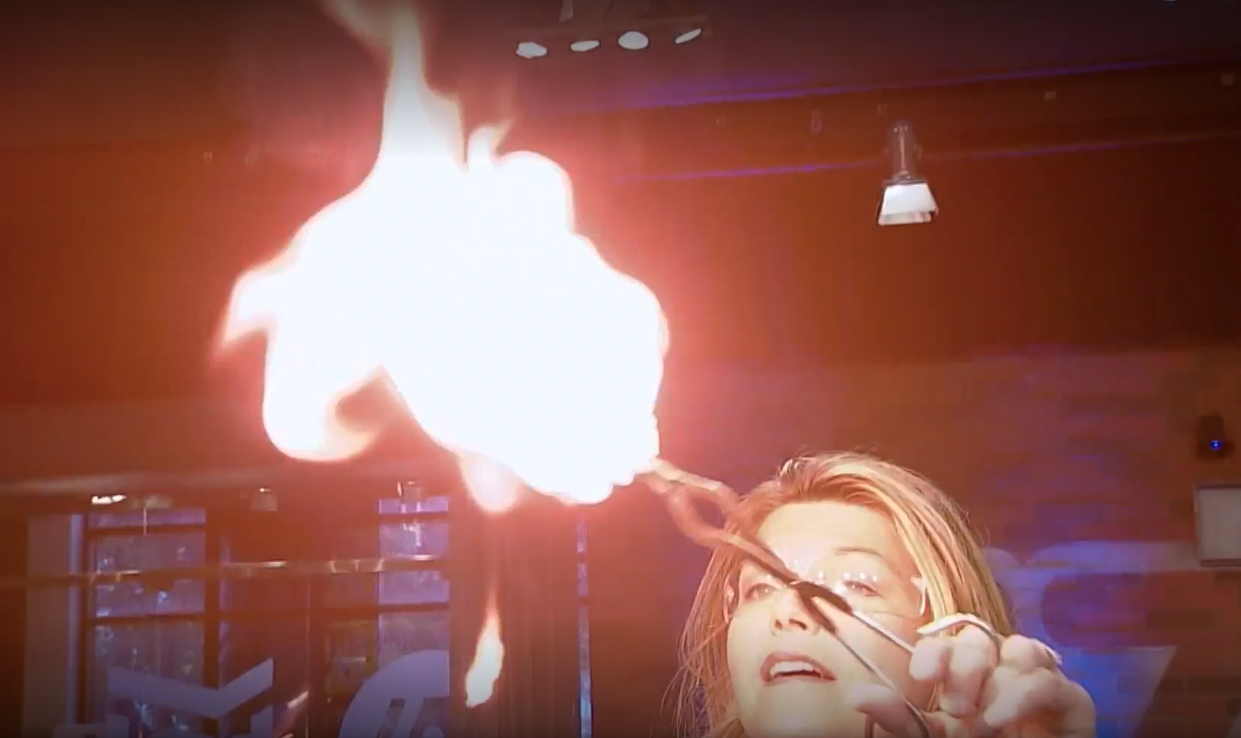 A woman with safety glasses holds something on fire in front of her.