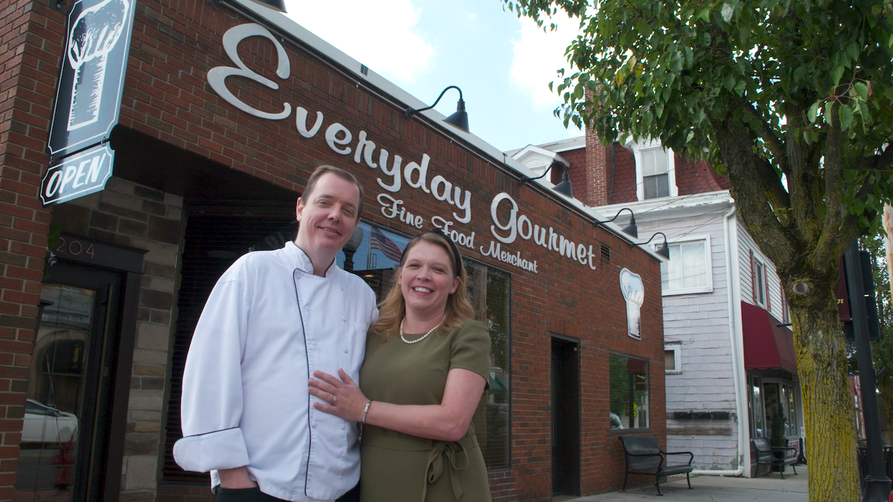 Woman in green shirt holding onto a man in a white chef coat standing in front of a brick building.