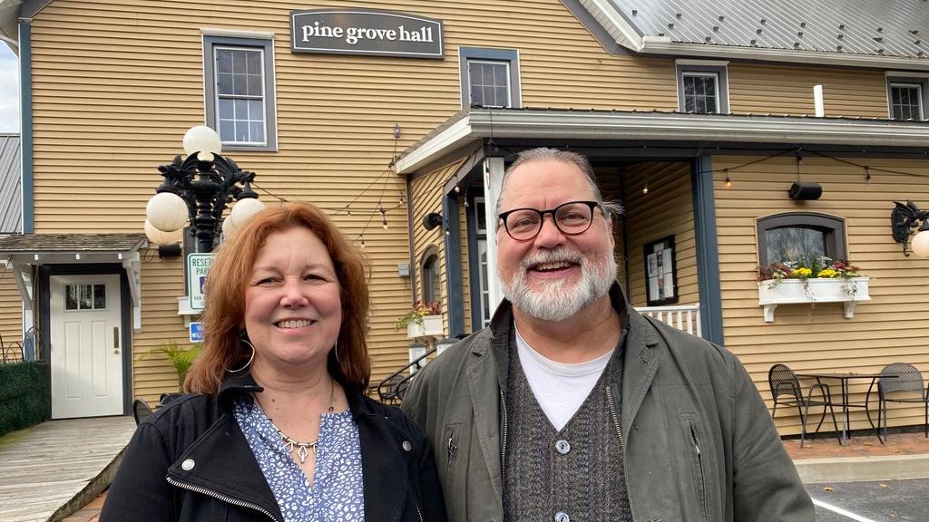 Woman with long red and brown hair standing along side a man with glasses and gray beard in front of a yellow building.