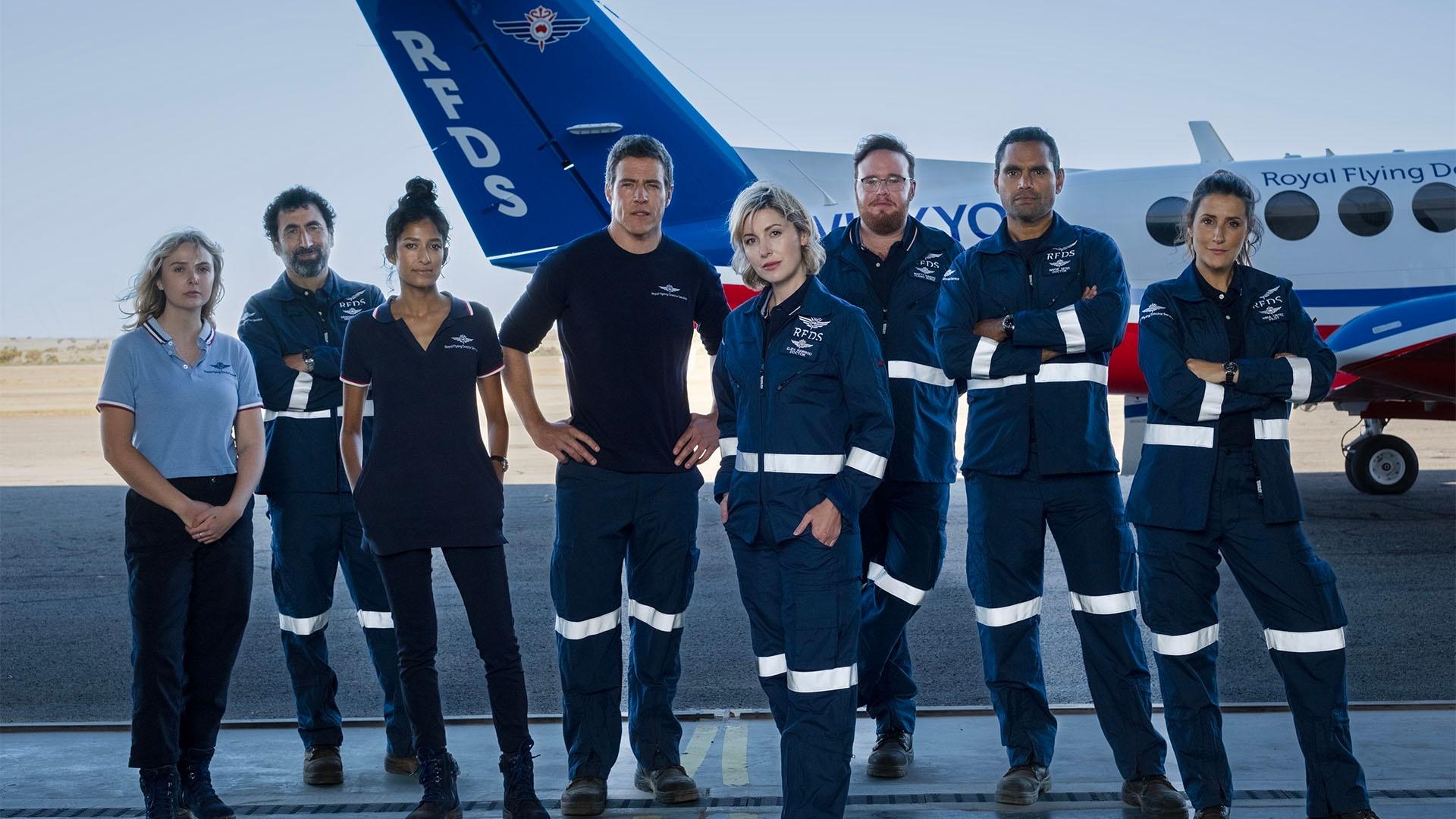Group of men and women in blue uniforms standing in a hanger with a plane in the background.