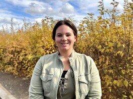 Woman in tan jacket standing in front of shrubs.