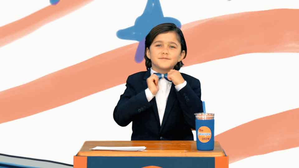 Kid wearing a suit and bowtie standing behind podium in front of a red and white striped background.