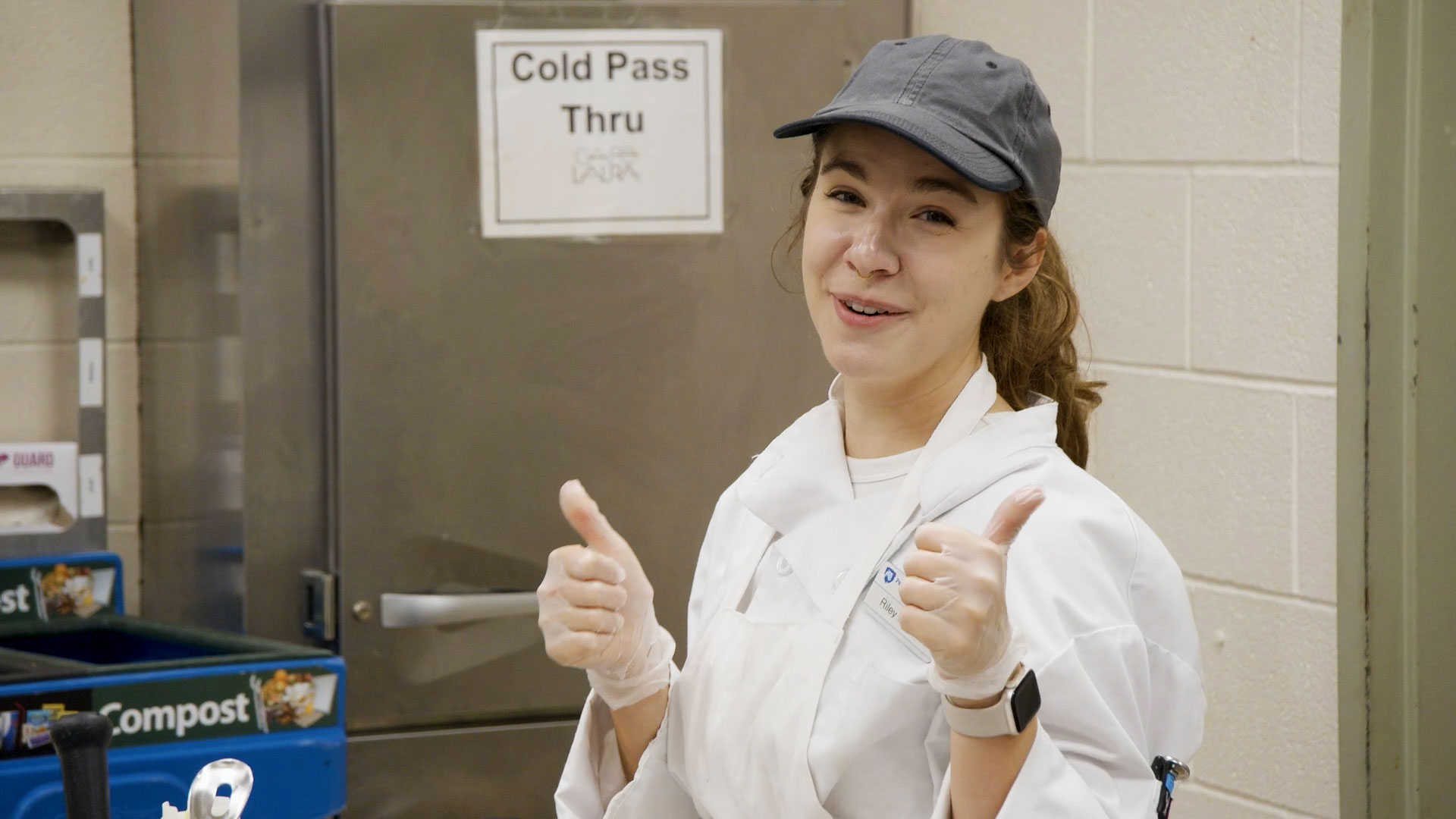Woman in a chef coat and blue cap smiling and giving thumbs up.