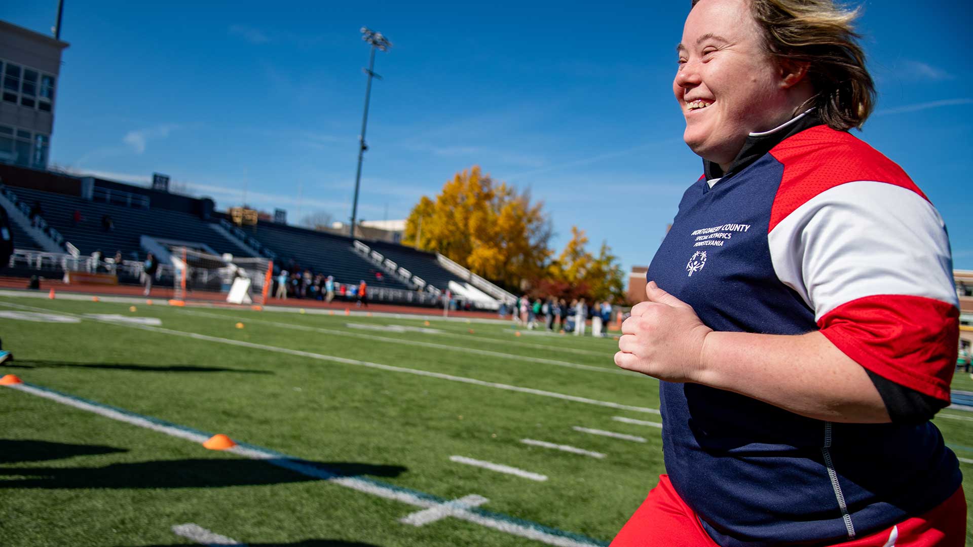 Female in blue jacket with red and white striped sleeves running across a football field.