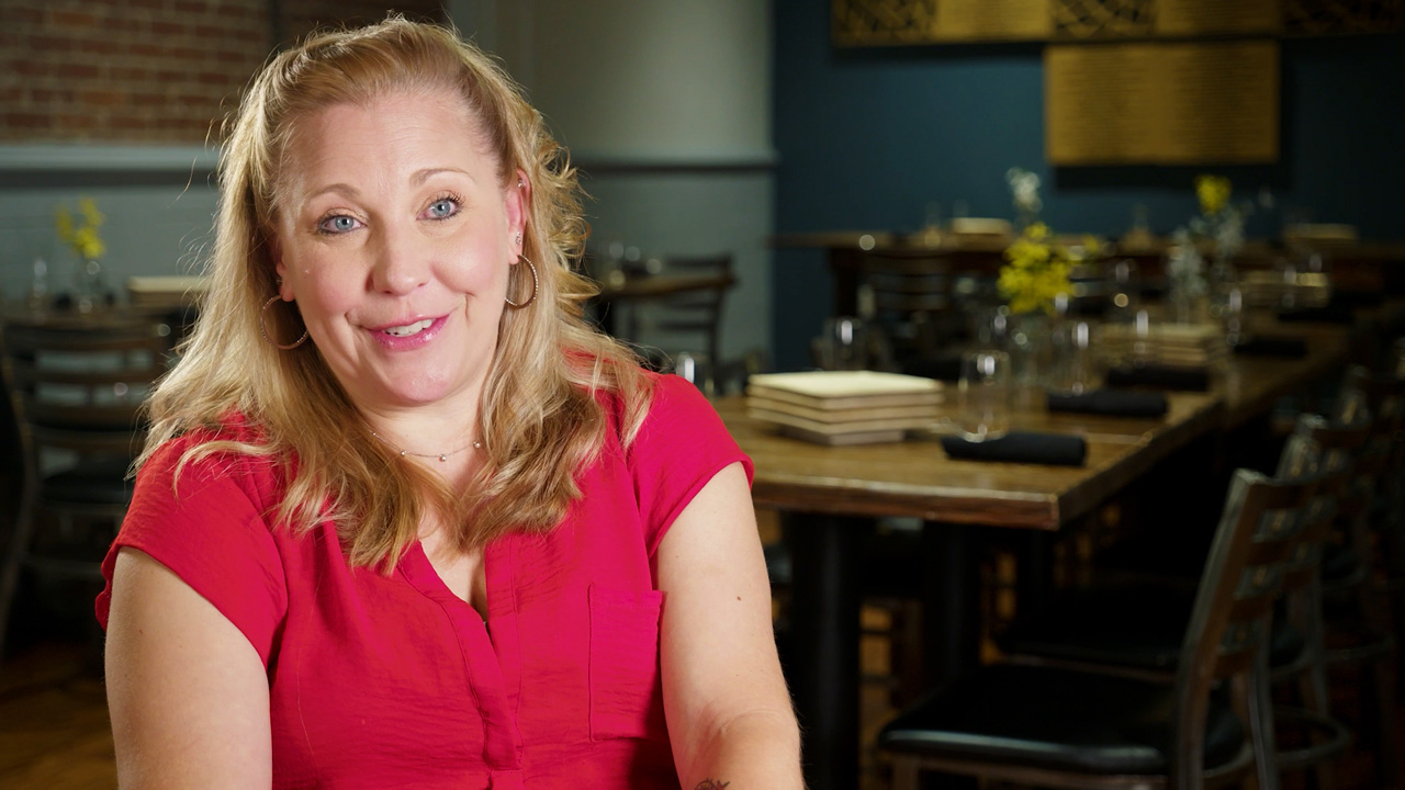 Woman with blonde hair and red shirt sitting with restaurant table in background.