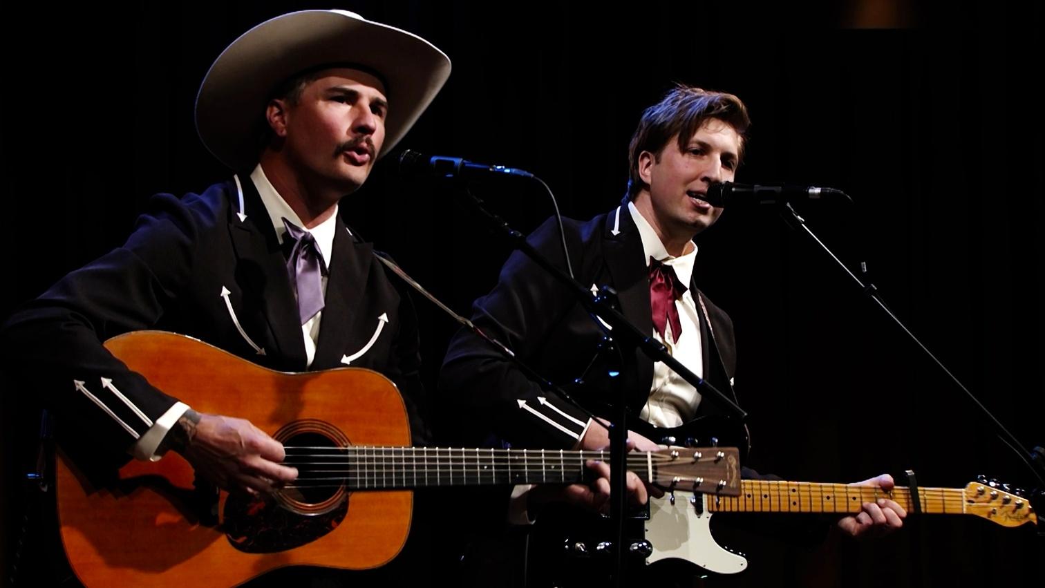 Man in cowboy hat plays guitar while singing into microphone.