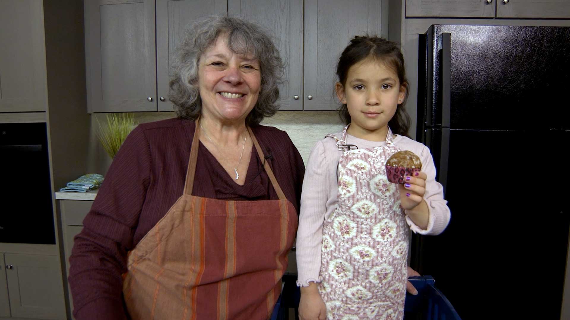 Young girl holding orange cranberry nut muffin, standing next to older woman in apron.