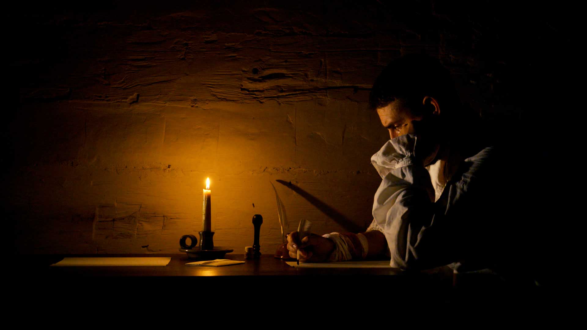 Man in colonial period clothing writing a letter with quill pen by candle light.
