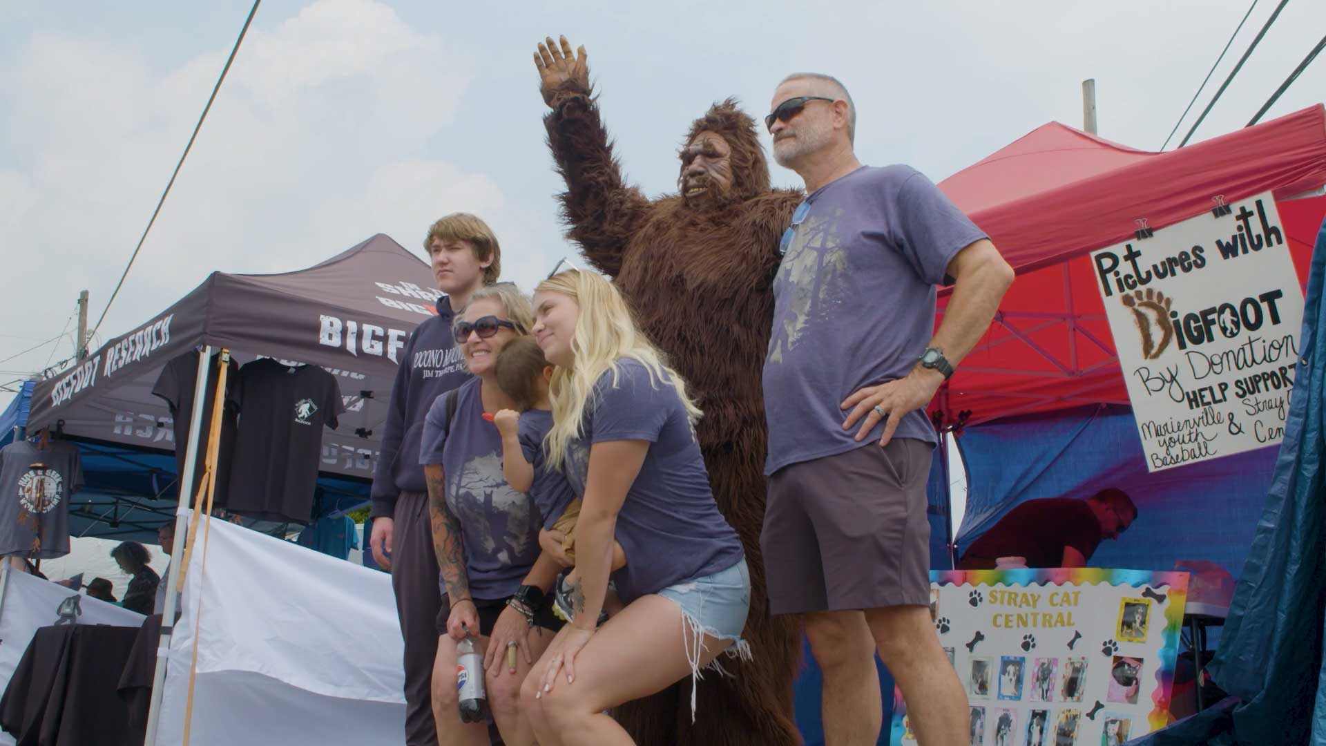 Men and women posing for a picture with a statue of Bigfoot at an outdoor festival.