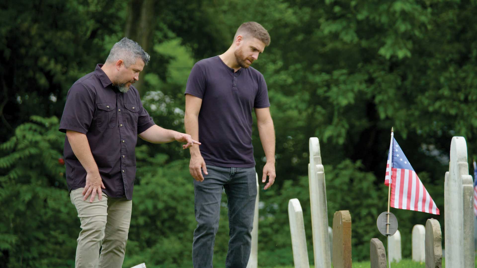 Two men looking down a headstones in a cemetary.