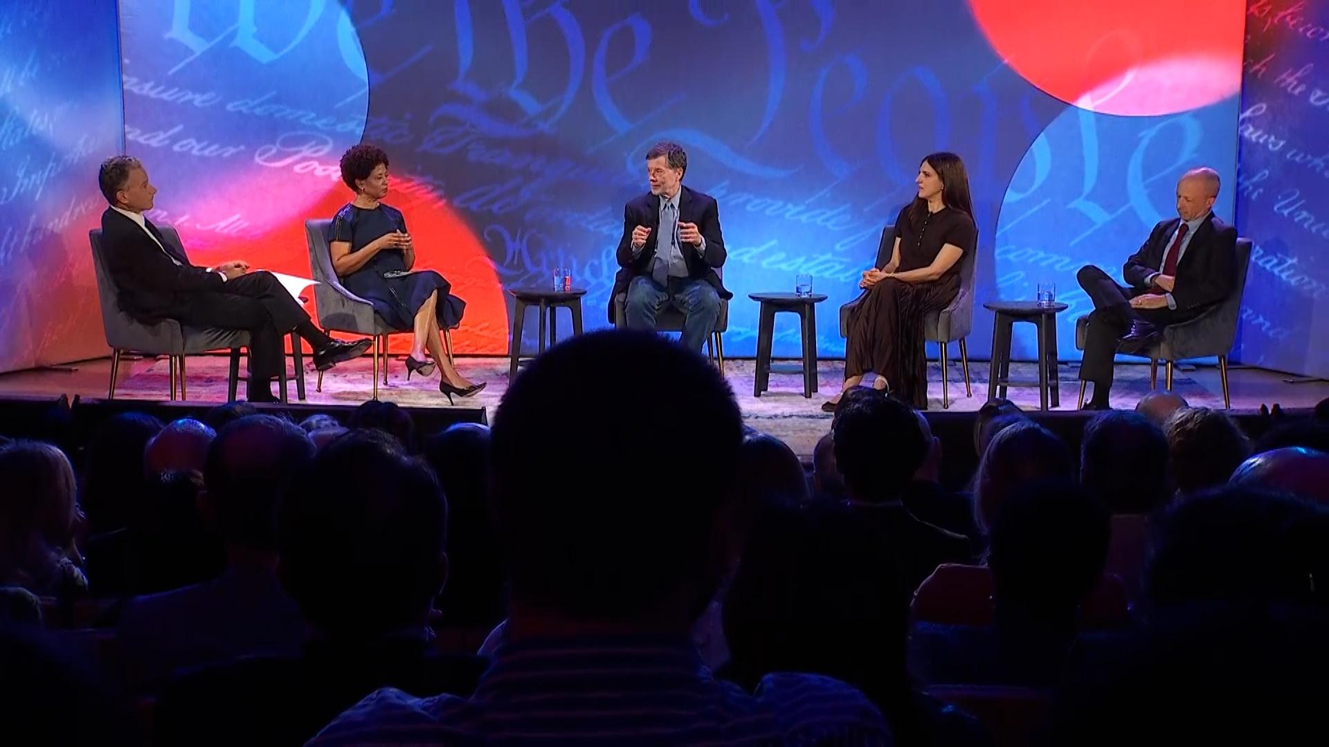 A panel of five ment and women on a stage with a patriotic background.