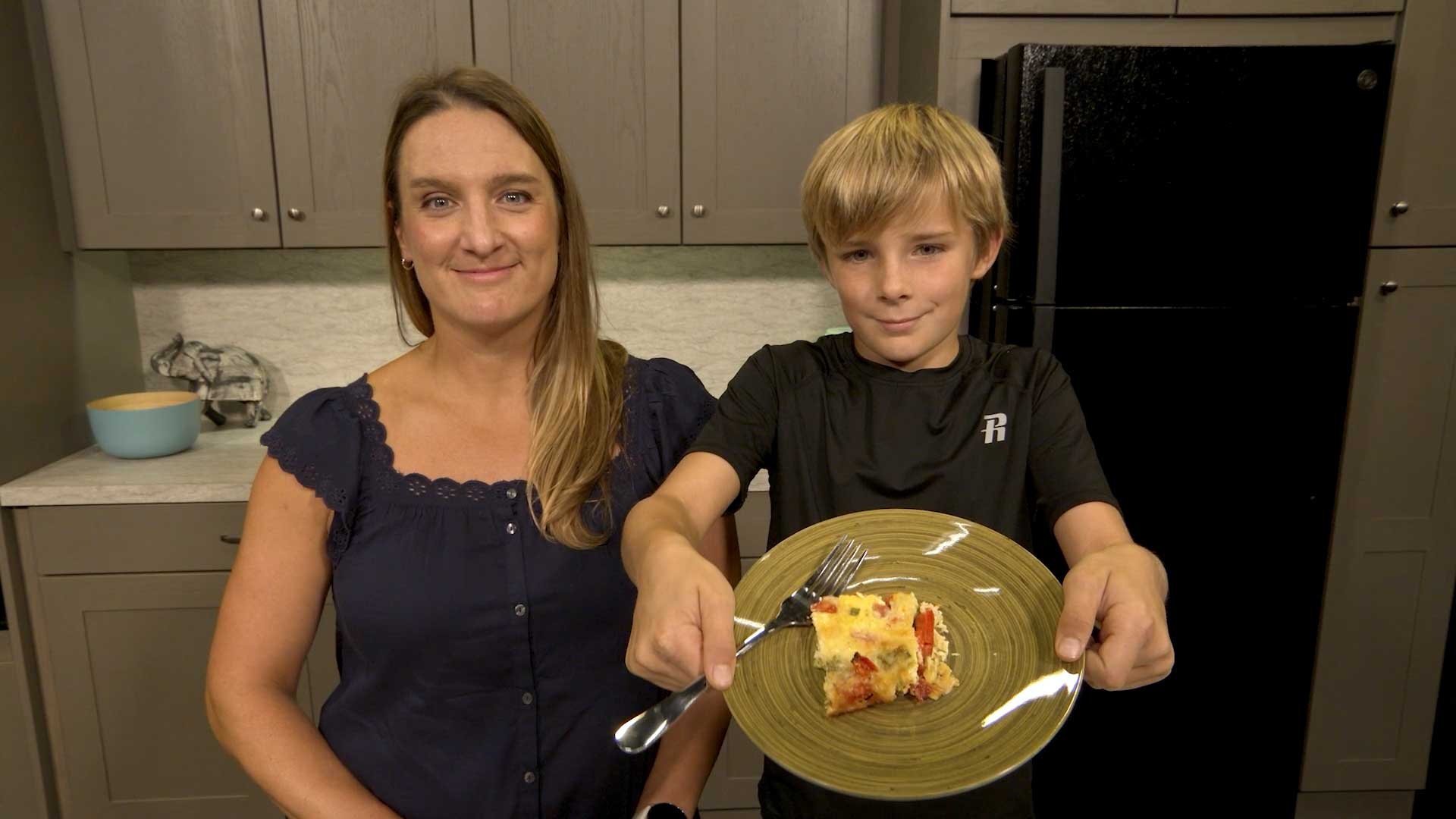 Woman standing next to teenage boy holding a plate with a piece of broccoli frittata.