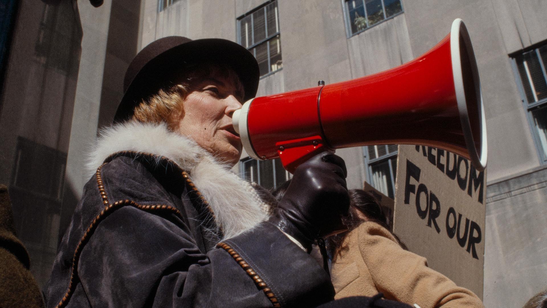 Woman in a bowler hat and winter coat speaking into a red megaphone.