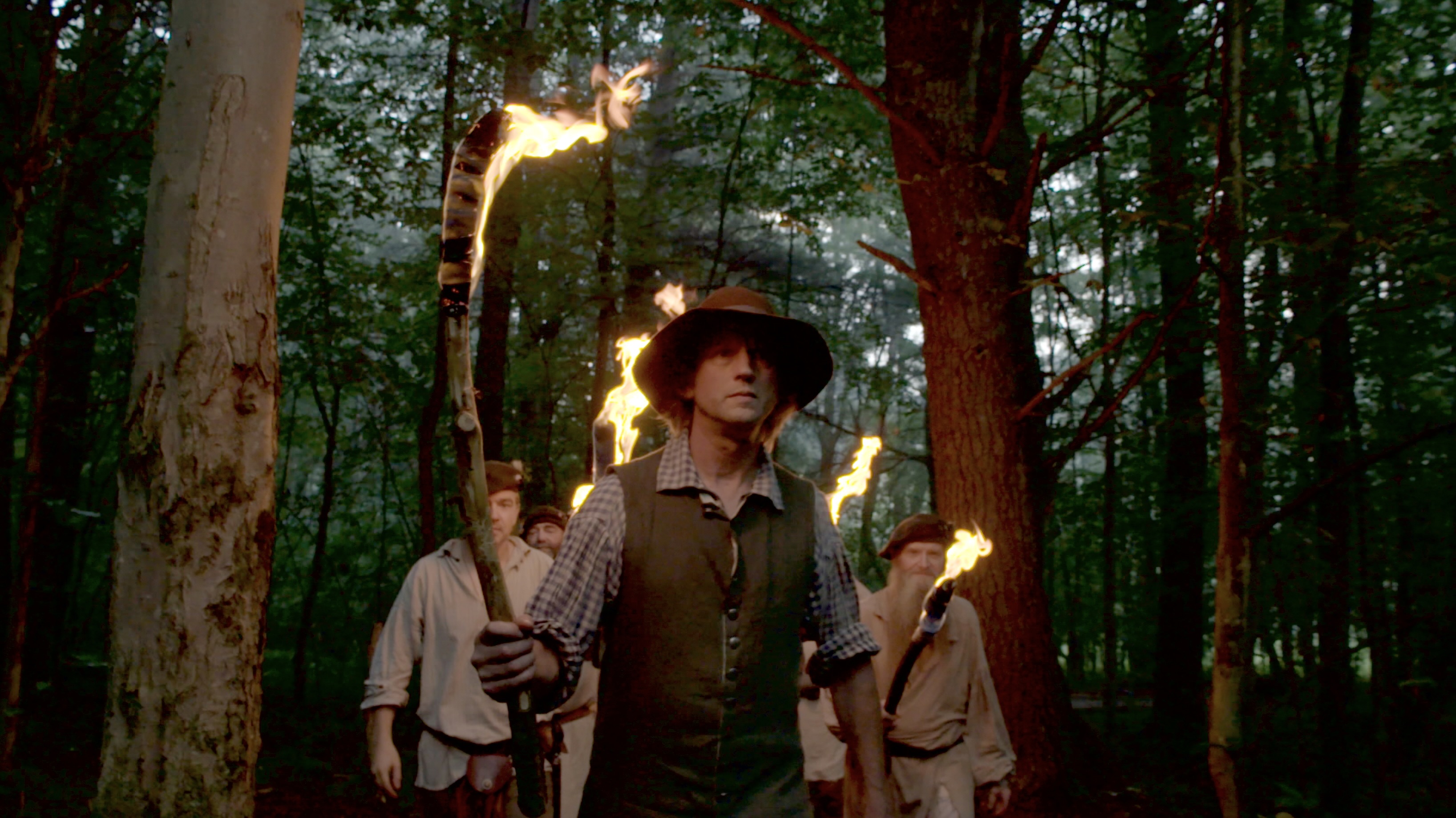 Historical figures passing through a wooded area holding torches.