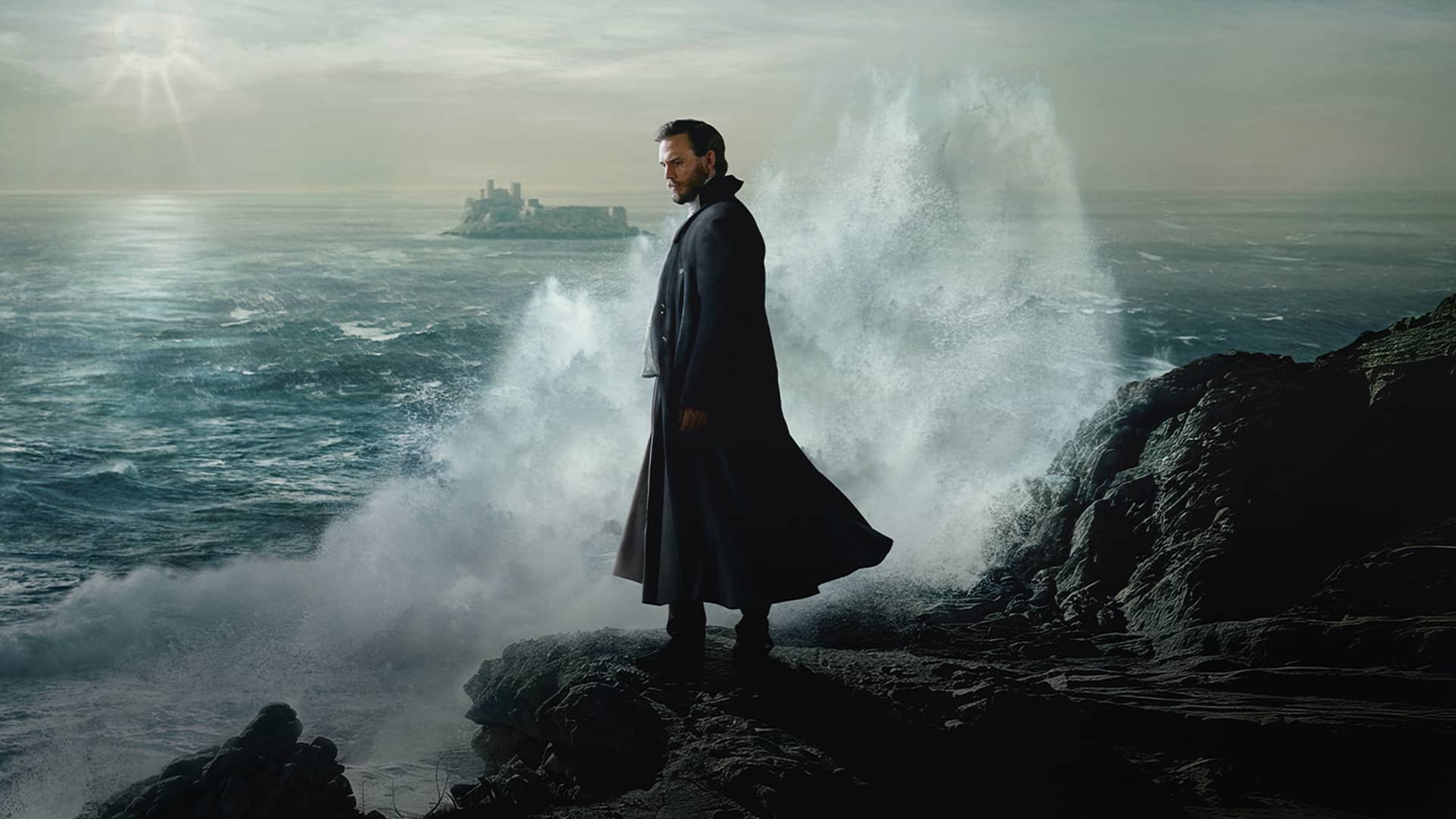 Man in long coat standing on rocks in front of the crashing waves of the ocean.