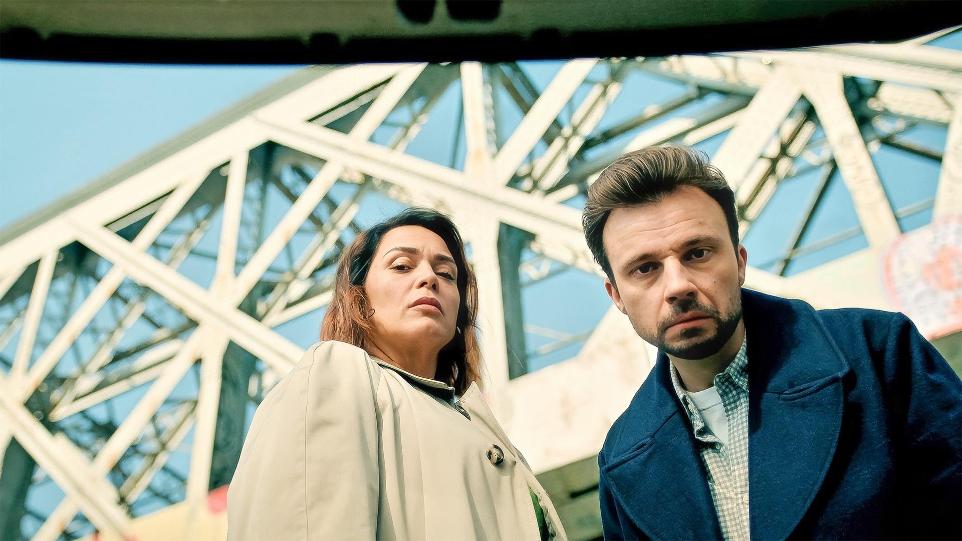 Man and woman looking down at the camera with a large bridge in the background.