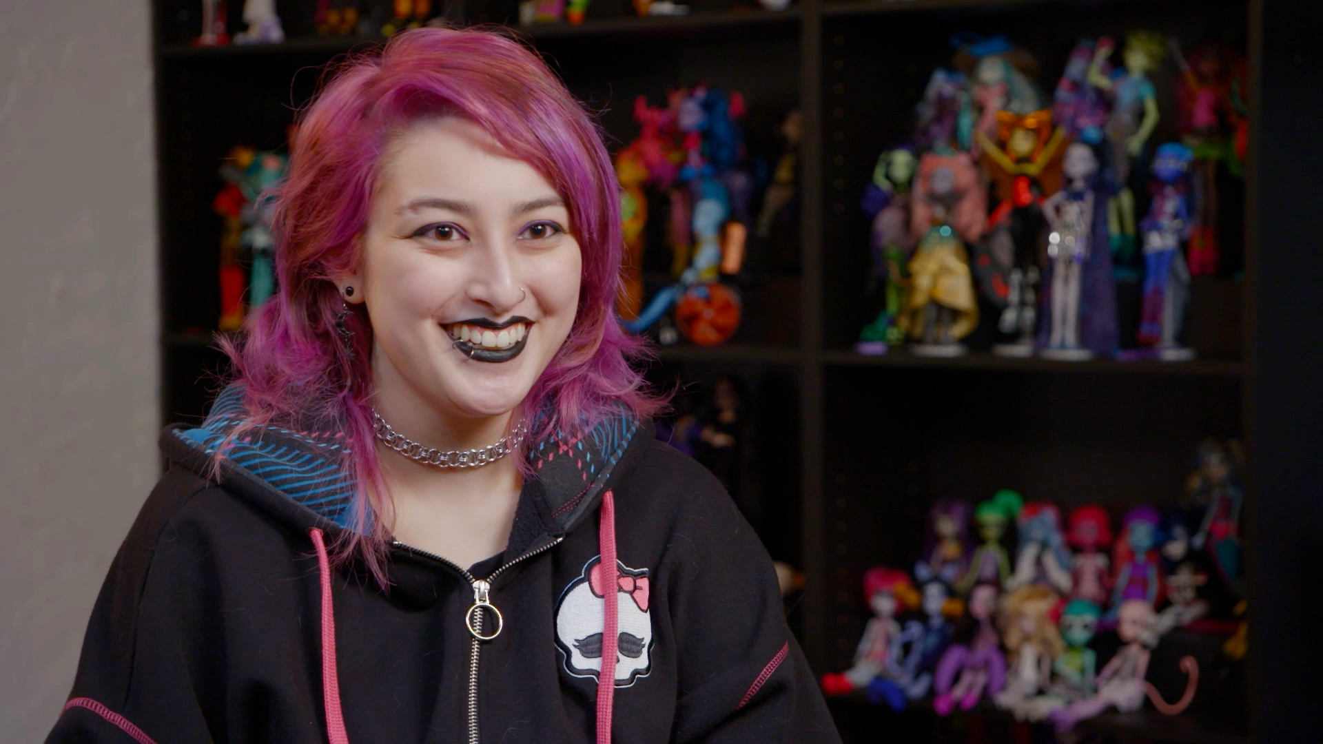 Violet Burney, wearing a black hooded sweatshirt and with pink hair, speaks in front of her collection of Monster High dolls.
