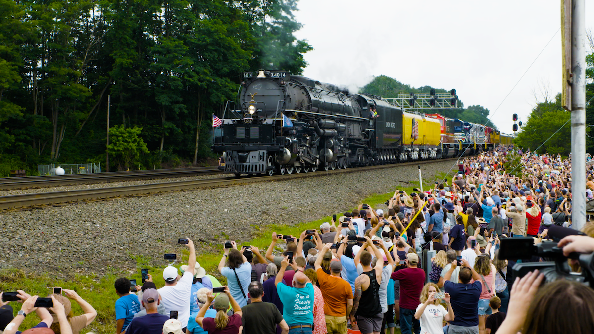 A large steam locomotive rolling down a train track in front of a large crowd of people.